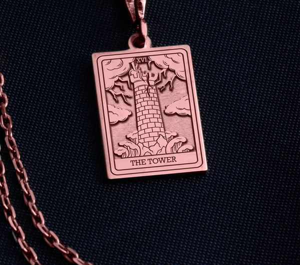 The Tower tarot card rose gold boho chic pendant necklace , depicted in a striking rose gold color features a tall, imposing structure being struck by highlighting the crumbling tower as it tilts precariously. Two figures are shown falling from the structure, symbolizing sudden upheaval or chaos. The background is often minimal, allowing the vivid rose gold tones to emphasize the tower's destruction and the sense of unexpected change or revelation it represents.
