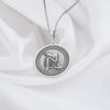 Sterling silver Buddha pendant necklace displaying a serene meditation scene. This detailed circular medallion captures Buddha in contemplative pose with head resting on hand, surrounded by an ornately engraved border. The pendant features meticulous craftsmanship with an oxidized finish highlighting the peaceful expression and flowing robes. Displayed on white silk fabric with a matching silver chain, this spiritual jewelry piece symbolizes tranquility, mindfulness