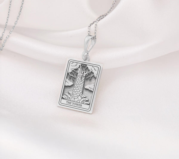 The Tower tarot card silver boho chic pendant necklace , depicted in a striking silver color features a tall, imposing structure being struck by highlighting the crumbling tower as it tilts precariously. Two figures are shown falling from the structure, symbolizing sudden upheaval or chaos. The background is often minimal, allowing the vivid silver tones to emphasize the tower's destruction and the sense of unexpected change or revelation it represents.