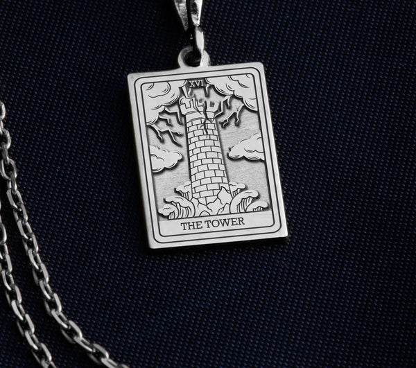 The Tower tarot card silver boho chic pendant necklace , depicted in a striking silver color features a tall, imposing structure being struck by highlighting the crumbling tower as it tilts precariously. Two figures are shown falling from the structure, symbolizing sudden upheaval or chaos. The background is often minimal, allowing the vivid silver tones to emphasize the tower's destruction and the sense of unexpected change or revelation it represents.