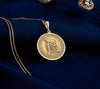 14K solid gold Buddha pendant necklace featuring a detailed medallion of Buddha in resting pose. This circular pendant showcases Buddha with head resting on hand in peaceful meditation, with intricate relief engravings and ornate border design. The pendant has an antique finish highlighting the serene details and hangs from a delicate gold chain against white silk fabric. This premium 14K gold spiritual jewelry piece represents peace, enlightenment
