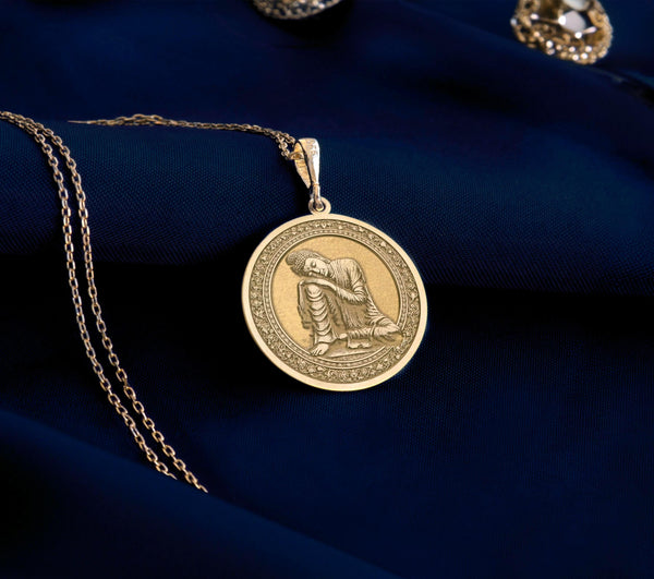 14K solid gold Buddha pendant necklace featuring a detailed medallion of Buddha in resting pose. This circular pendant showcases Buddha with head resting on hand in peaceful meditation, with intricate relief engravings and ornate border design. The pendant has an antique finish highlighting the serene details and hangs from a delicate gold chain against white silk fabric. This premium 14K gold spiritual jewelry piece represents peace, enlightenment
