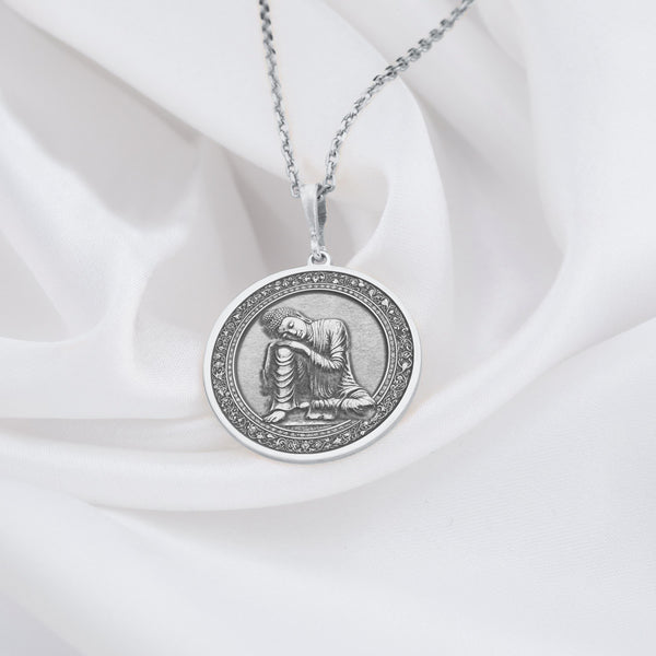 Sterling silver Buddha pendant necklace displaying a serene meditation scene. This detailed circular medallion captures Buddha in contemplative pose with head resting on hand, surrounded by an ornately engraved border. The pendant features meticulous craftsmanship with an oxidized finish highlighting the peaceful expression and flowing robes. Displayed on white silk fabric with a matching silver chain, this spiritual jewelry piece symbolizes tranquility, mindfulness