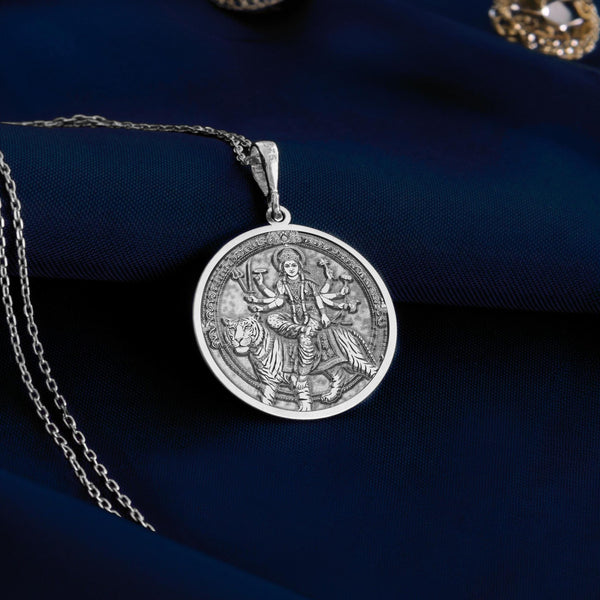 Sterling silver Durga pendant necklace showcasing the revered Hindu goddess seated on her tiger vahana. This detailed circular medallion captures Durga with multiple arms holding symbolic weapons and objects. The pendant features meticulous engravings with an oxidized finish to highlight the intricate details of the goddess and her tiger mount. Displayed on white silk fabric with a matching silver chain, this spiritual jewelry piece symbolizes feminine strength, protection against evil