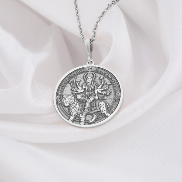 Sterling silver Durga pendant necklace showcasing the revered Hindu goddess seated on her tiger vahana. This detailed circular medallion captures Durga with multiple arms holding symbolic weapons and objects. The pendant features meticulous engravings with an oxidized finish to highlight the intricate details of the goddess and her tiger mount. Displayed on white silk fabric with a matching silver chain, this spiritual jewelry piece symbolizes feminine strength, protection against evil