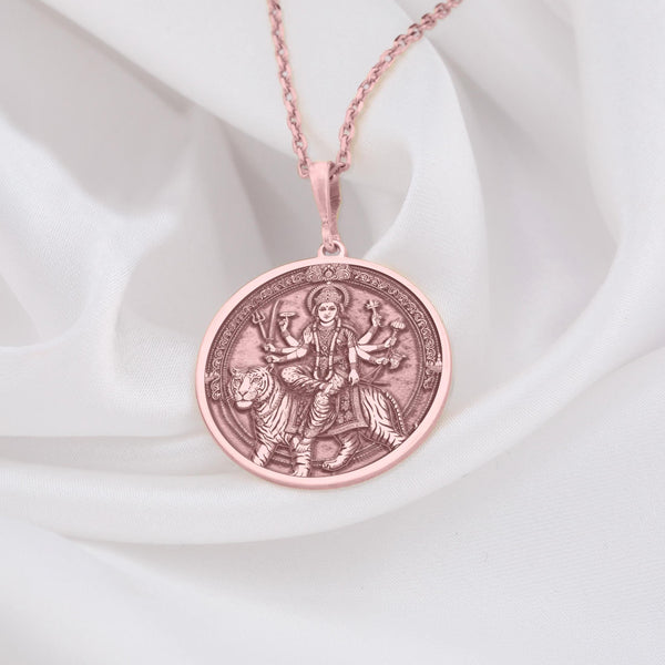 Rose gold plated Durga pendant necklace displaying the Hindu goddess of power and protection riding her tiger. This circular medallion features beautifully detailed engravings showing multi-armed Durga holding traditional weapons and symbolic items. The warm rose gold plating creates an elegant contrast with the antique finish of the engraved details, all suspended from a matching chain against soft white fabric. This spiritual jewelry piece celebrates divine feminine energy,