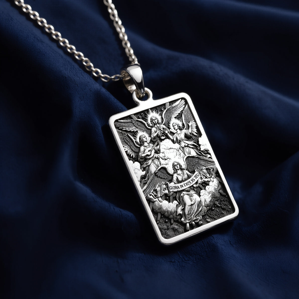 Rectangular sterling silver pendant necklace featuring a detailed relief engraving of four angels playing musical instruments with the Latin inscription "Gloria in Excelsis Deo," suspended from a polished cable chain on deep blue fabric.