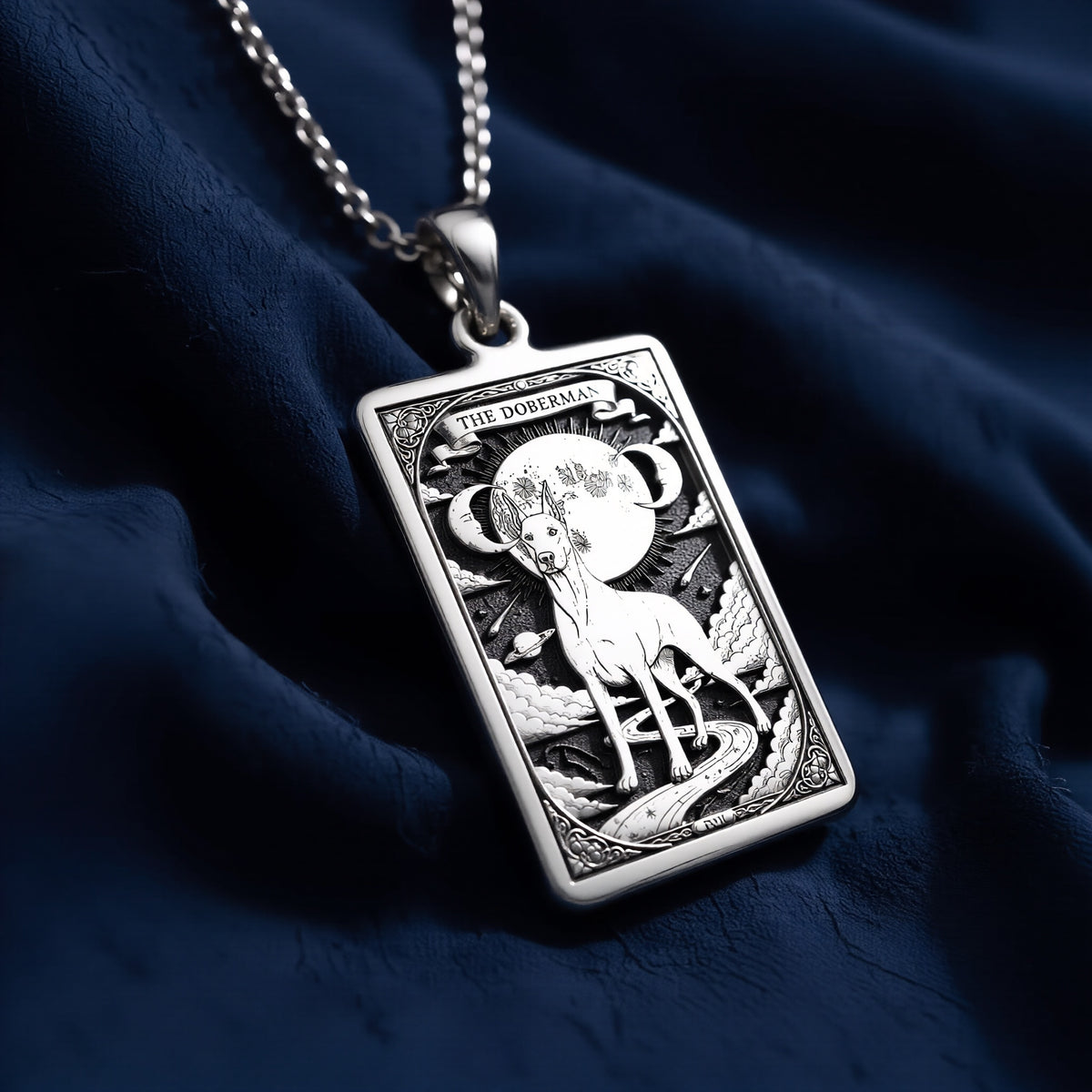 Rectangular silver pendant necklace featuring a detailed engraving of a Doberman standing before a celestial moon with stars, clouds, and cosmic symbols, suspended from a fine link chain on dark blue fabric.