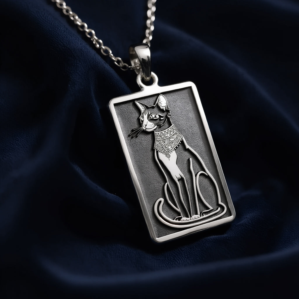 Rectangular silver pendant necklace featuring a detailed engraving of Bastet, the Egyptian goddess depicted as a seated cat with ornate collar, suspended from a fine link chain against dark fabric.
