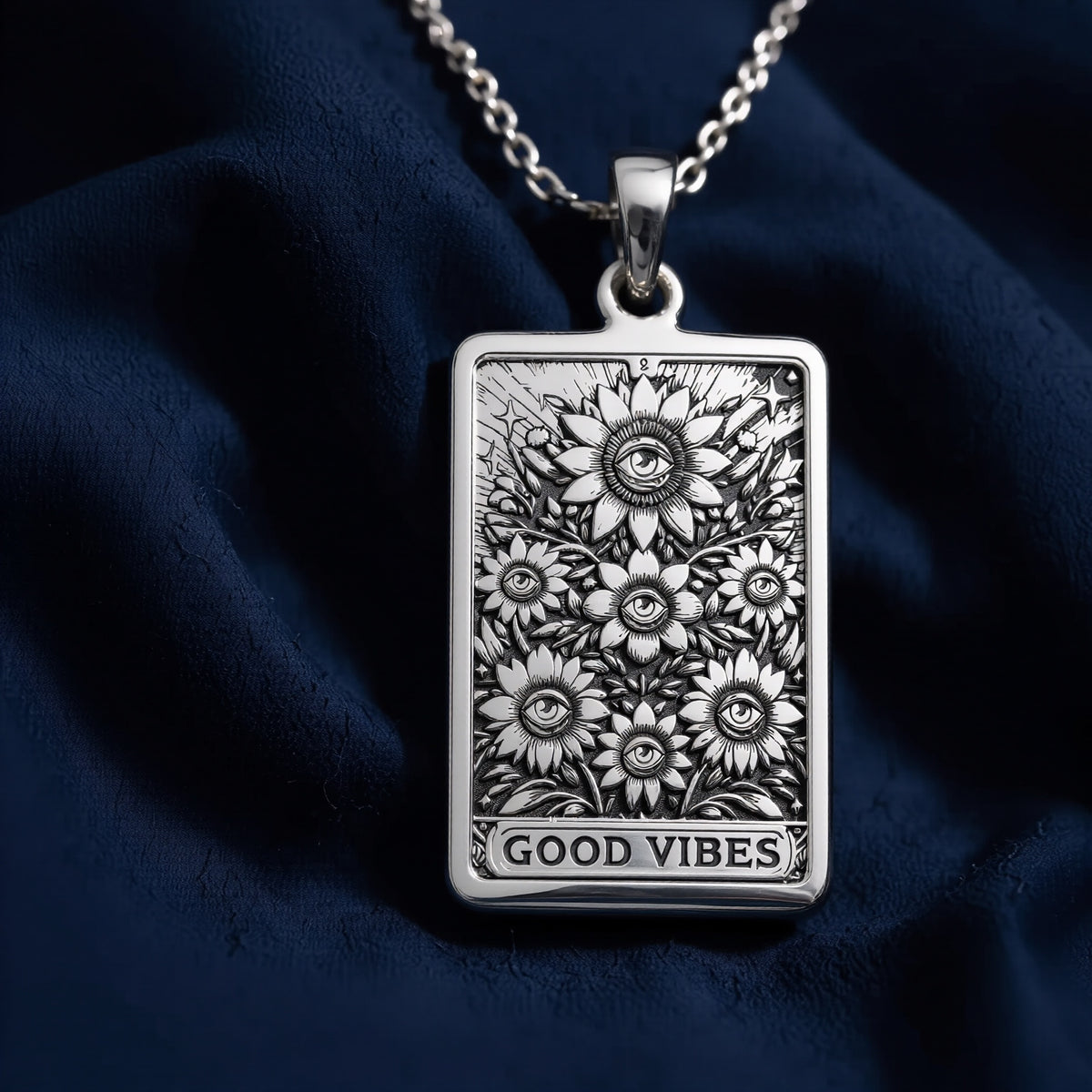 Silver rectangular pendant showing a detailed sunflower and floral pattern with a border and the text "GOOD VIBES". The pendant is worn on a silver chain, resting on black velvet fabric surrounded by loose gemstones.