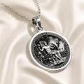 Silver circular medallion pendant displaying an ancient mythological scene of a male figure. The pendant is attached to a silver chain and rests on a dark blue fabric background with white flowers.