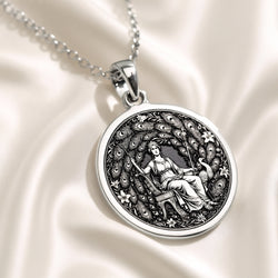 Silver circular pendant with an engraved classical motif of a seated female figure surrounded by detailed foliage. This pendant is attached to a silver chain and displayed on a dark blue surface.
