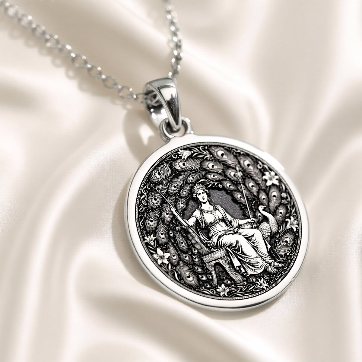 Silver circular pendant with an engraved classical motif of a seated female figure surrounded by detailed foliage. This pendant is attached to a silver chain and displayed on a dark blue surface.