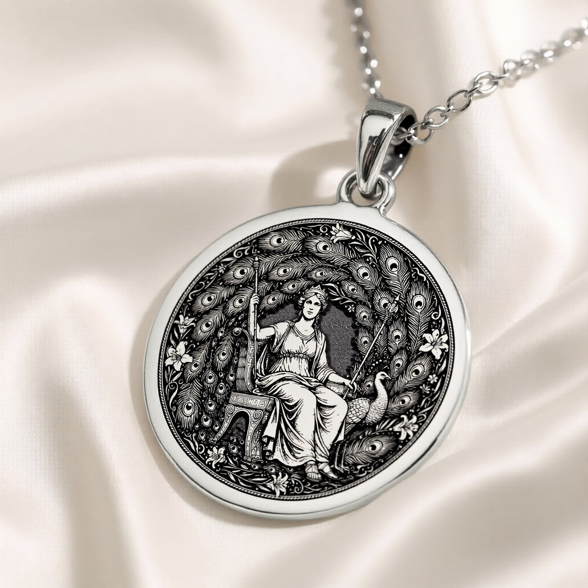 Silver circular pendant featuring an engraved classical design of a seated female figure within a decorative wreath of leaves. The pendant hangs from a silver chain over a blue fabric background.