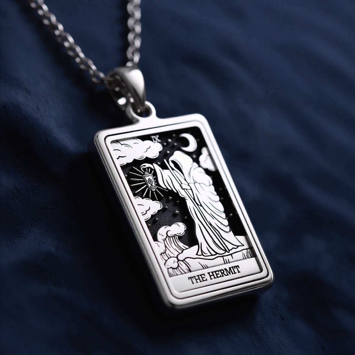 Rectangular sterling silver pendant featuring engraved The Hermit tarot card with robed figure holding lantern, crescent moon, clouds, stars, Roman numeral IX, and “THE HERMIT” inscription at base, suspended from a polished link chain on dark blue fabric background.