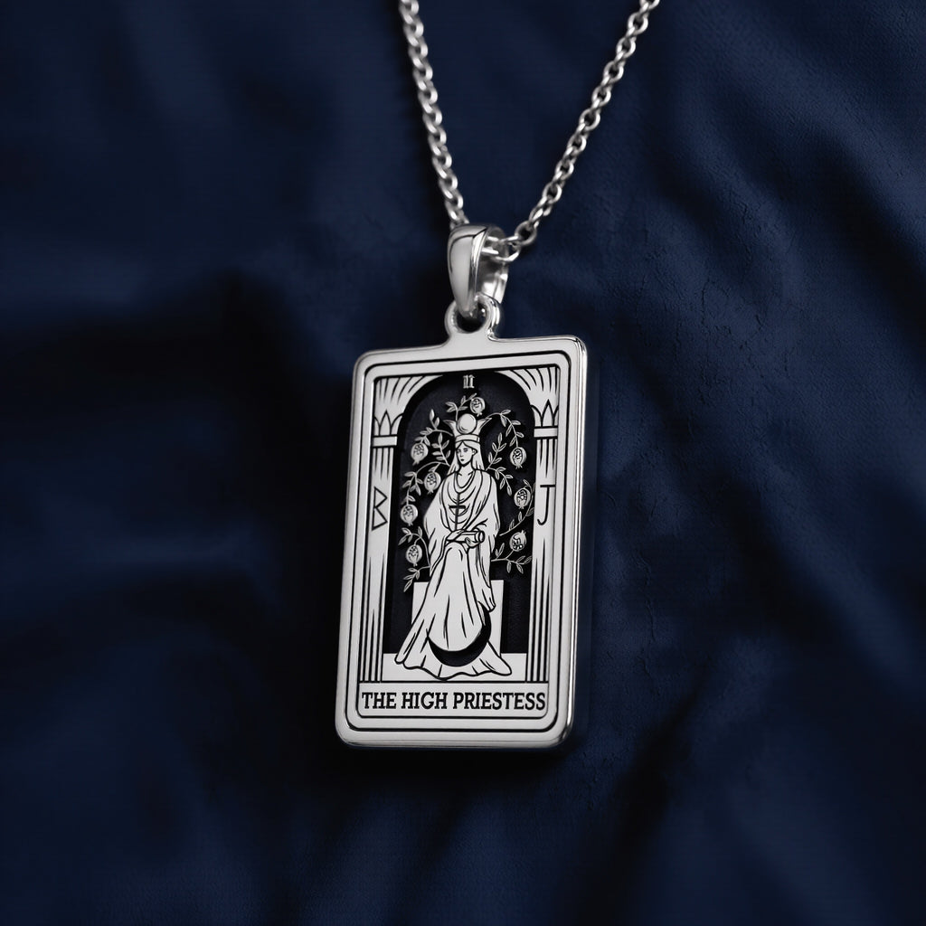 Rectangular sterling silver pendant necklace featuring an engraved High Priestess tarot card design with seated figure, flanking columns, floral motifs, and “THE HIGH PRIESTESS” inscription at base, suspended from a fine link chain against navy fabric.