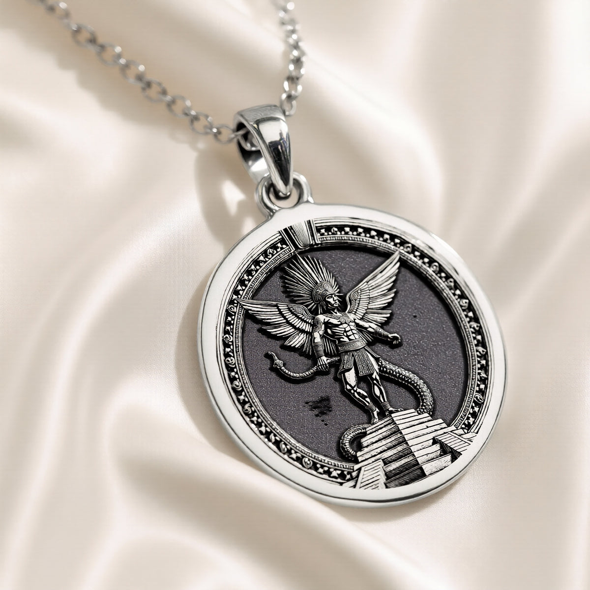 Silver circular pendant featuring an intricate Aztec winged deity design resting on a serpentine structure. The pendant is attached to a silver chain