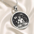 Silver plated circular pendant featuring a figure seated in a meditative pose within a decorative border. The pendant is attached to a silver chain and rests on a dark blue fabric background.