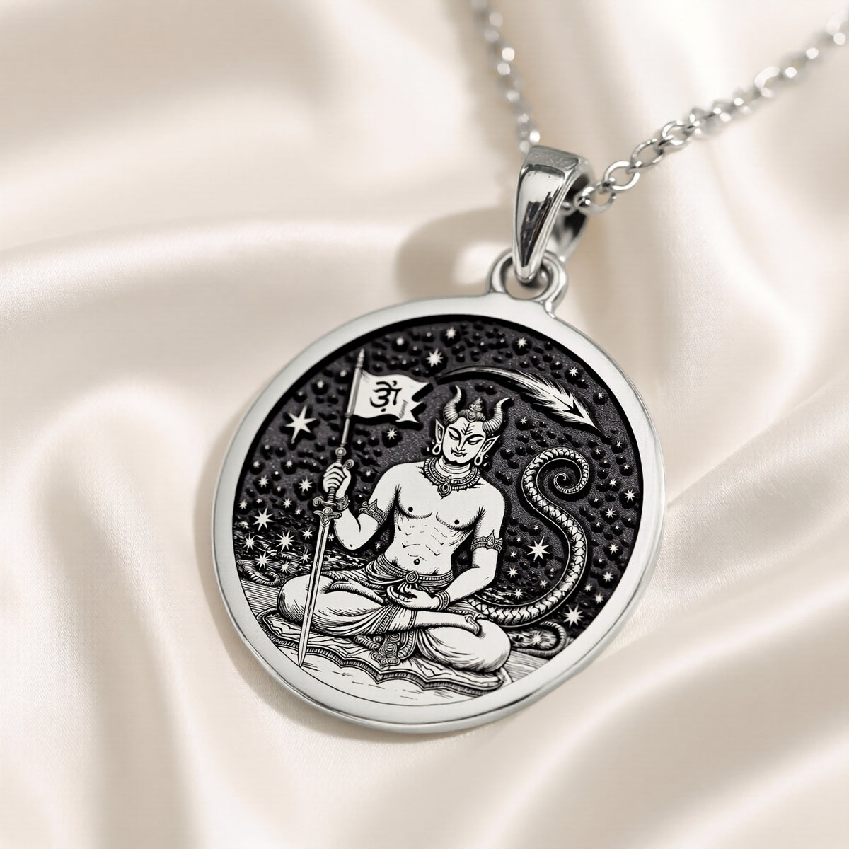 Silver pendant with a circular design featuring a stylized figure in a meditative pose surrounded by swirling patterns. The surface has a matte finish and is shown from a slightly elevated angle against a dark blue background.