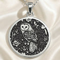 Silver pendant featuring an intricate design of an owl surrounded by detailed floral and leaf patterns. The pendant is attached to a silver chain and rests on draped fabric