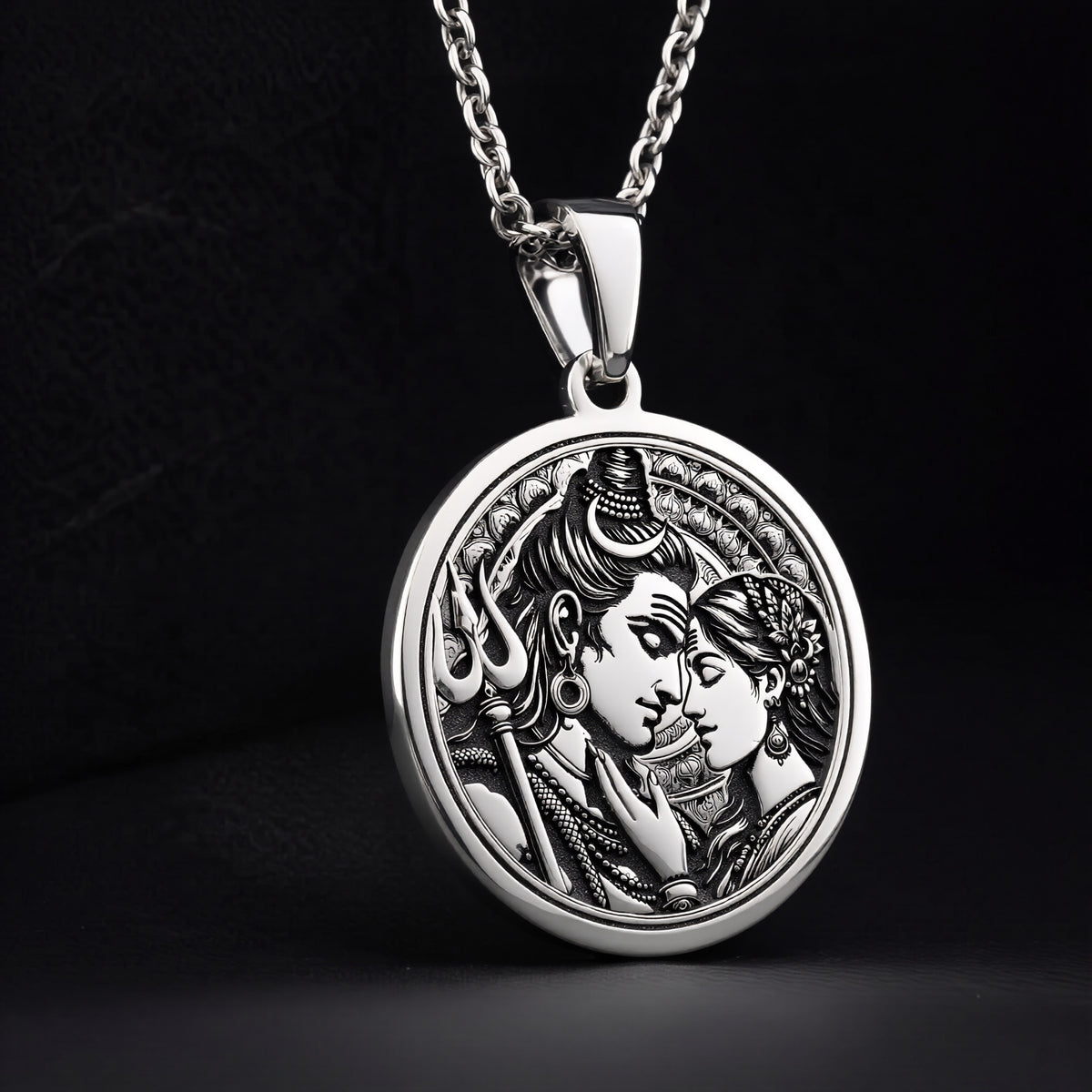 Silver toned circular pendant showing a detailed relief carving of Shiva and Parvati. This necklace features a silver chain and is displayed against a dark blue fabric background.