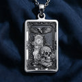 Silver rectangular pendant featuring an intricate engraved design of a skull, clock, and eagle. This piece is displayed on a silver chain surrounded by loose gemstones and dark textile.