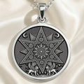Silver pendant featuring an intricate star design with geometric patterns and stylized figures at the bottom. The pendant is attached to a chain and rests on draped fabric and a dark surface.
