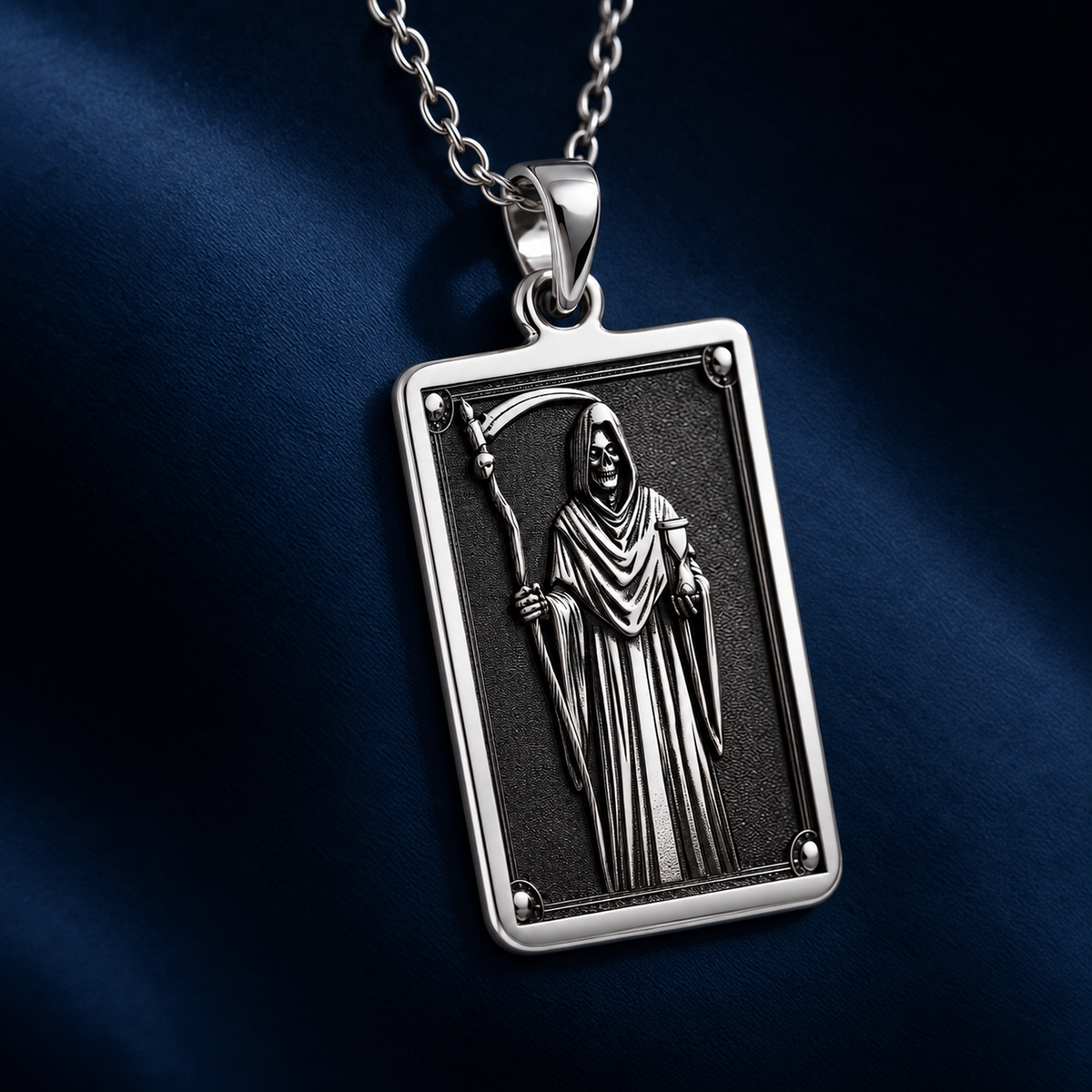 Silver pendant necklace showing a framed design of a hooded figure holding a scythe and skulls. The item rests on black fabric surrounded by various cut, faceted gemstones.