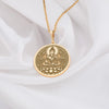 Buddha figure seated in meditative lotus pose, upon a blooming lotus flower base. The rich, traditional yellow gold finish of the quality plating highlights the detailed craftsmanship of the central deity and the surrounding ornate floral mandala-style patterns filling the circular frame. This spiritual Buddhist necklace symbolizes peace and enlightenment, shown hanging from a chain against a soft white fabric background with natural dried grasses. Perfect yoga or mindfulness jewelry gift