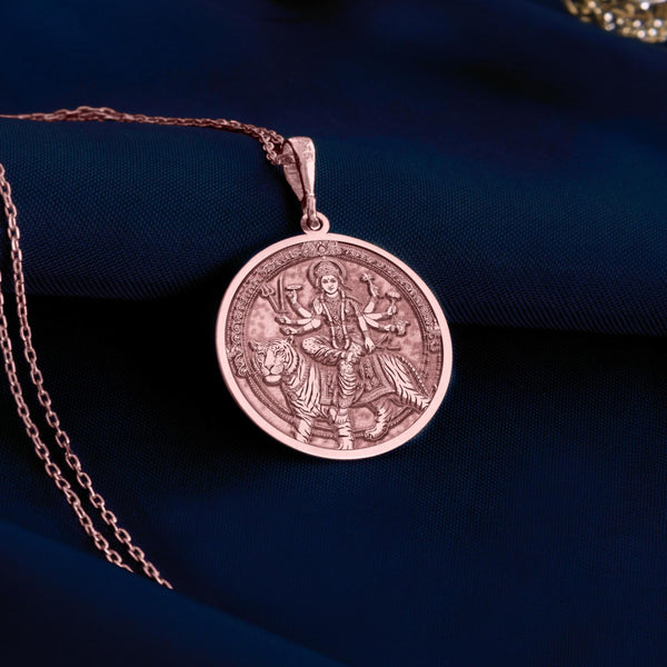 Rose gold plated Durga pendant necklace displaying the Hindu goddess of power and protection riding her tiger. This circular medallion features beautifully detailed engravings showing multi-armed Durga holding traditional weapons and symbolic items. The warm rose gold plating creates an elegant contrast with the antique finish of the engraved details, all suspended from a matching chain against soft white fabric. This spiritual jewelry piece celebrates divine feminine energy,
