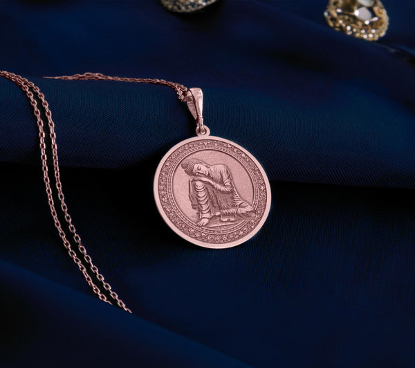 Rose gold plated Buddha pendant necklace showcasing a peaceful meditation scene. This circular medallion features beautifully detailed engravings of Buddha in restful contemplation, head gently resting on hand. The warm rose gold plating creates an elegant contrast with the antique finish of the intricate border details, all suspended from a matching chain against soft white fabric.