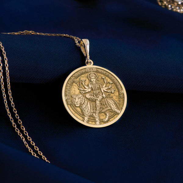 14K solid gold Durga pendant necklace featuring the powerful Hindu goddess riding a tiger. This detailed medallion shows Durga with multiple arms holding traditional weapons and symbolic items. The circular pendant has an antique finish with intricate engravings and hangs from a delicate gold chain against white silk fabric. This premium 14K gold religious jewelry piece represents divine feminine power, protection, and strength for spiritual devotees and fine jewelry collectors.