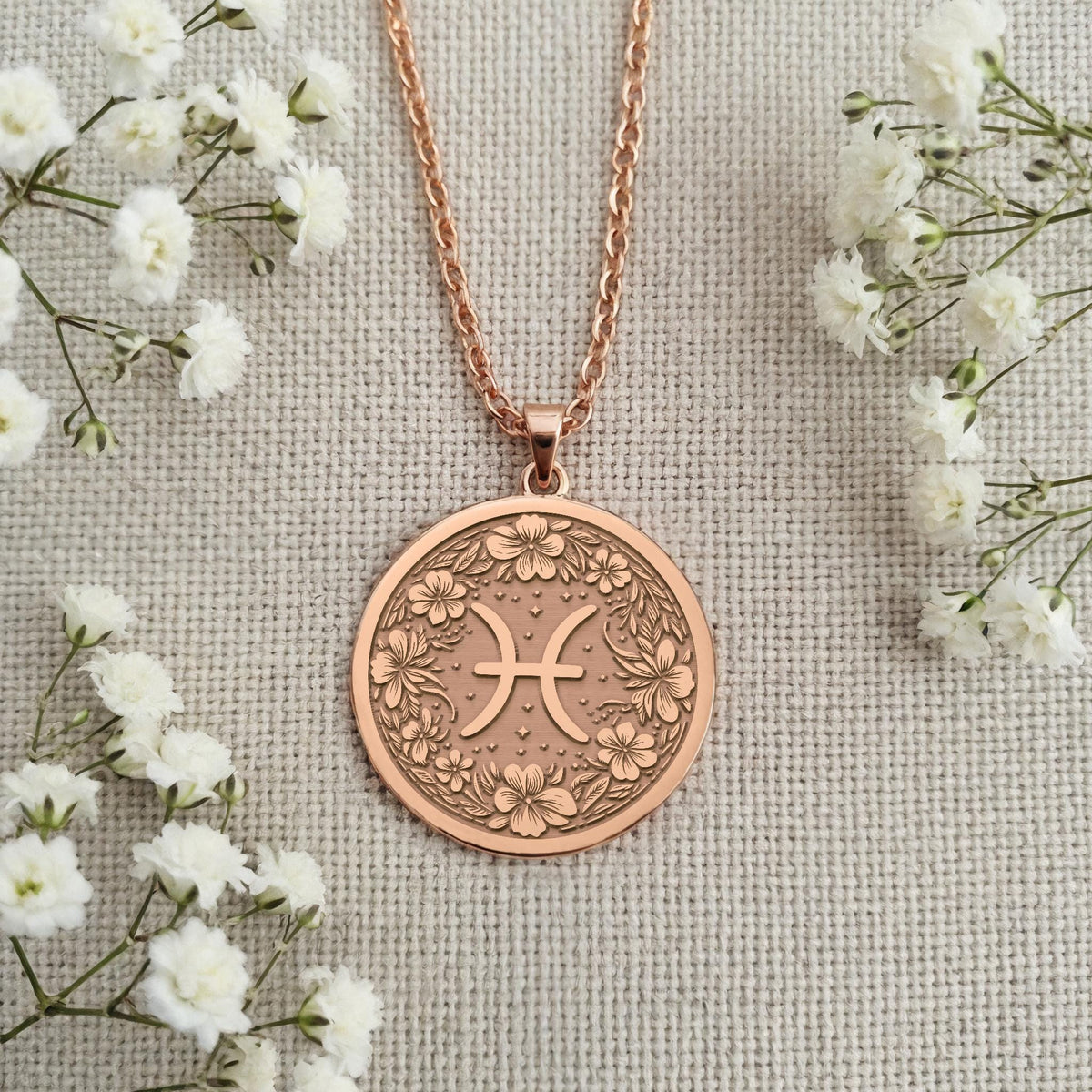 Engravable Rose Gold Plated Pisces Pendant on a textured surface. The floral horoscope symbol medallion is a perfect personalized gift for a March or February birthday. Customizable jewelry.