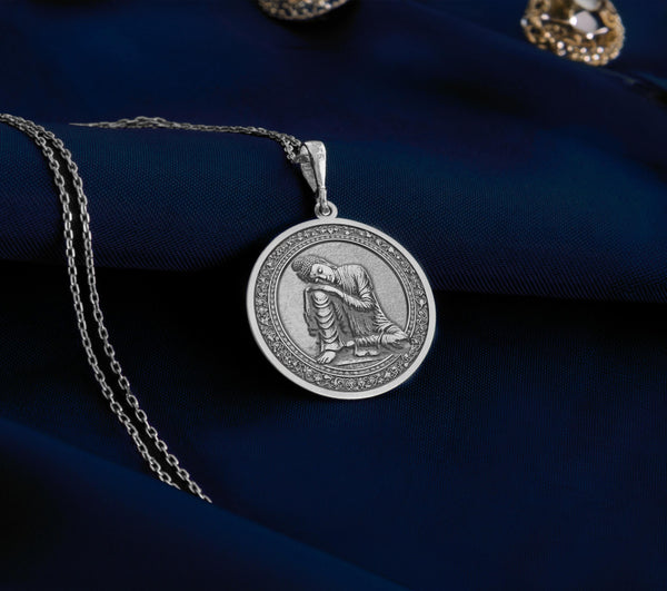 Sterling silver Buddha pendant necklace displaying a serene meditation scene. This detailed circular medallion captures Buddha in contemplative pose with head resting on hand, surrounded by an ornately engraved border. The pendant features meticulous craftsmanship with an oxidized finish highlighting the peaceful expression and flowing robes. Displayed on white silk fabric with a matching silver chain, this spiritual jewelry piece symbolizes tranquility, mindfulness
