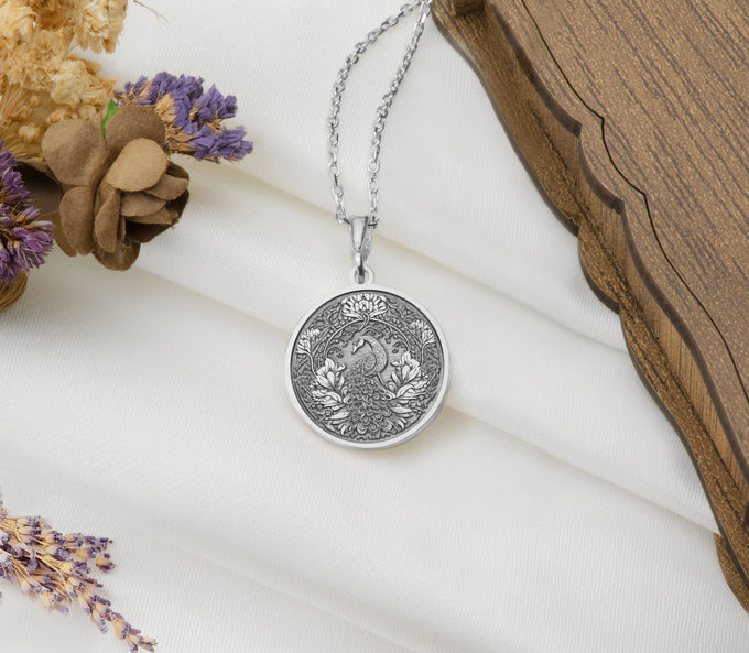 Silver colored circular pendant featuring an intricate embossed design of a peacock surrounded by detailed foliage. The pendant is attached to a silver-toned chain and displayed on a textured surface.
