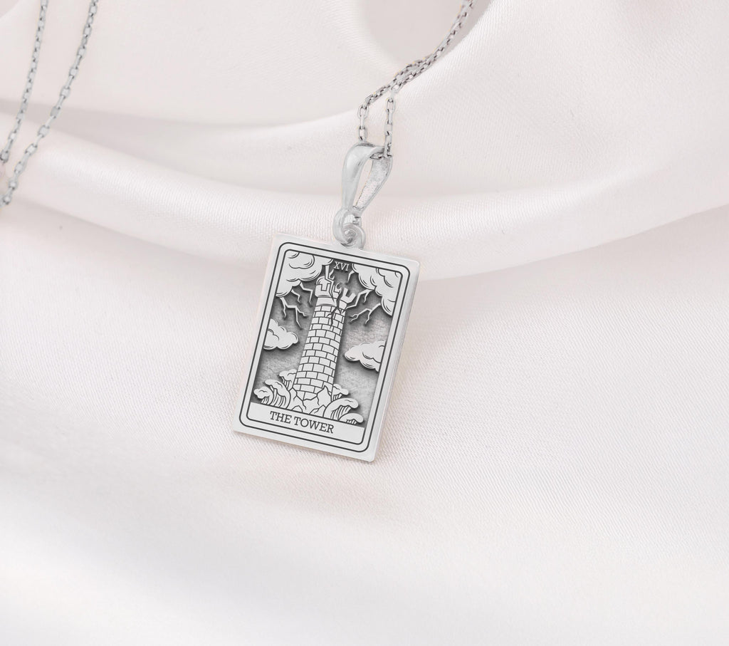 The Tower tarot card silver boho chic pendant necklace , depicted in a striking silver color features a tall, imposing structure being struck by highlighting the crumbling tower as it tilts precariously. Two figures are shown falling from the structure, symbolizing sudden upheaval or chaos. The background is often minimal, allowing the vivid silver tones to emphasize the tower's destruction and the sense of unexpected change or revelation it represents.