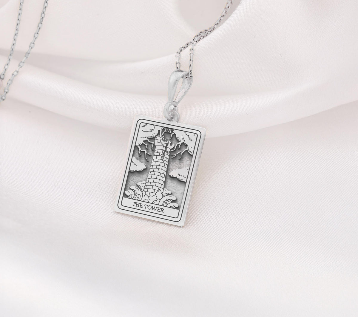 The Tower tarot card silver boho chic pendant necklace , depicted in a striking silver color features a tall, imposing structure being struck by highlighting the crumbling tower as it tilts precariously. Two figures are shown falling from the structure, symbolizing sudden upheaval or chaos. The background is often minimal, allowing the vivid silver tones to emphasize the tower's destruction and the sense of unexpected change or revelation it represents.