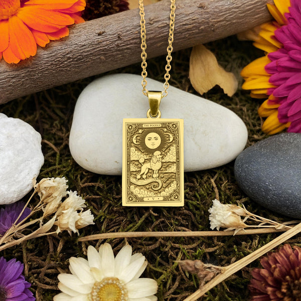 A gold-colored pendant necklace with a rectangular pendant featuring a tarot card design, surrounded by various natural elements such as rocks, flowers, and moss.
