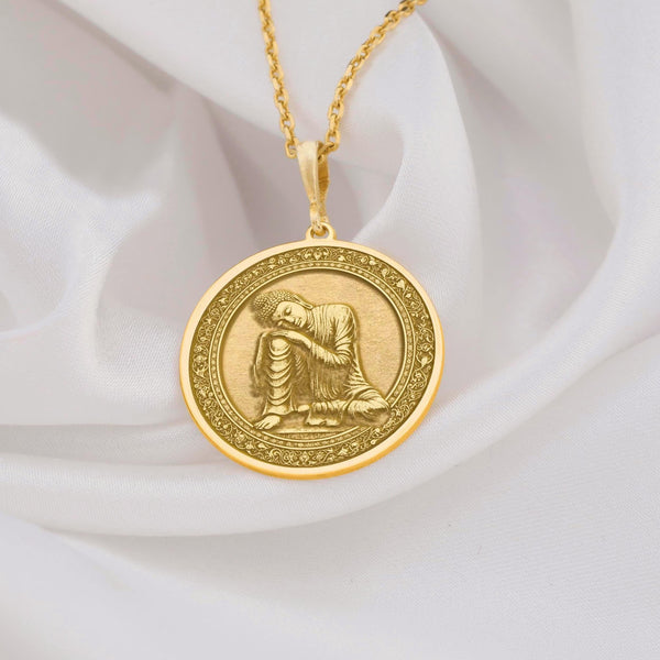14K solid gold Buddha pendant necklace featuring a detailed medallion of Buddha in resting pose. This circular pendant showcases Buddha with head resting on hand in peaceful meditation, with intricate relief engravings and ornate border design. The pendant has an antique finish highlighting the serene details and hangs from a delicate gold chain against white silk fabric. This premium 14K gold spiritual jewelry piece represents peace, enlightenment,