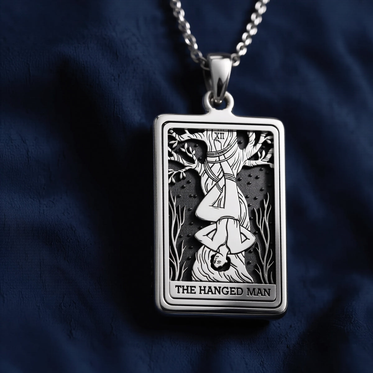 Rectangular sterling silver pendant featuring an engraved depiction of The Hanged Man tarot card with Roman numeral XII, suspended on a chain against dark fabric.