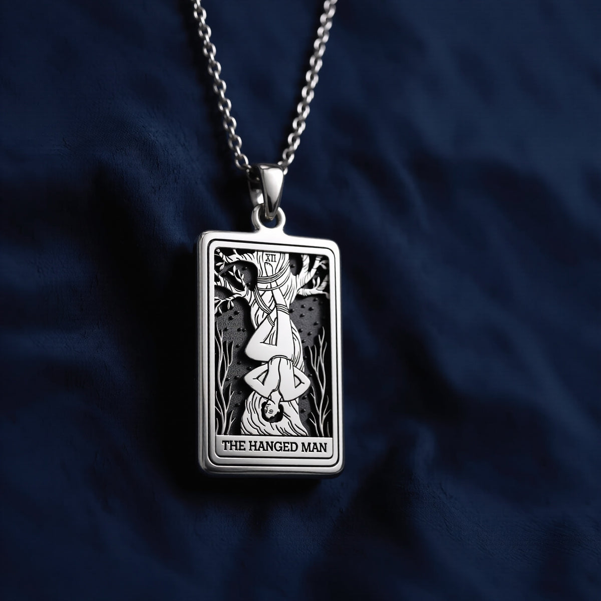 Rectangular sterling silver pendant featuring a detailed engraving of The Hanged Man tarot card with halo, suspended from a polished chain bail against deep blue fabric.