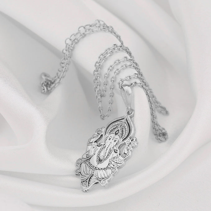 Silver toned pendant depicting the Ganesha motif with intricate detailing and ornate framing. The pendant is connected to a silver chain and rests on a white fabric background.