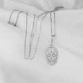 A silver toned pendant necklace laid out flat with an ornate skull design featuring scrollwork and a cross detail. A length of chain is visible alongside the pendant.