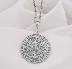 Silver pendant with an Aztec Sun Stone Calendar Medallion design. The circular pendant features intricate geometric patterns and depictions of ancient symbols in a detailed relief style. It is shown from the front, hanging from a silver chain.