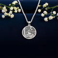 Silver circular pendant featuring an engraved scene of a woman interacting with sheep and a building in a landscape setting. The design is detailed and features classical figures. It is attached to a silver chain.
