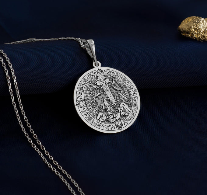 Silver circular pendant featuring an intricate relief design of religious figures framed by ornate borders. The pendant hangs from a silver-toned chain.