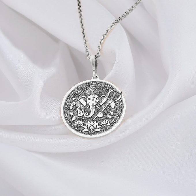 Silver circular pendant featuring an embossed design of the elephant-headed deity Ganesha with intricate detailing. The pendant hangs from a silver chain.