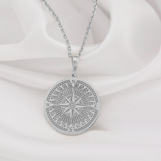 A silver-toned pendant necklace showcasing a detailed compass design within a circular frame, featuring intricate geometric patterns. The pendant is attached to a silver chain and rests on white fabric.