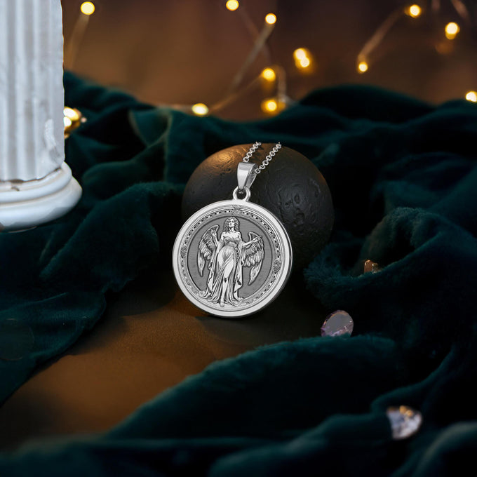 Silver plated circular pendant with an engraved winged female figure design. The pendant hangs from a silver chain and rests on dark fabric illuminated by string lights.