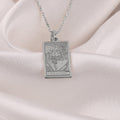 Silver pendant necklace featuring a rectangular design with an engraved depiction of two figures embracing, set against a smooth surface finish. The chain is visible, and the piece is displayed against a soft fabric background.